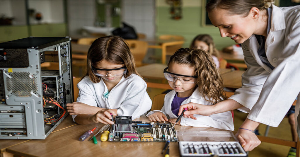 Girls in STEM - 2 Young Girls Working on Computer Hardware with Female Teacher, all wearing Lab Coats and Glasses